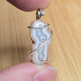 A close-up of a handmade pendant featuring a polished agate slice with natural brown and white banding, wrapped in a silver spiral wire, held between two fingers against a light wood background.