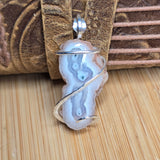 A silver-wrapped pendant featuring a polished agate slice with pale blue, gray, and white banding, displayed on a bamboo surface with a brown leather strap in the background.