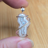 A close-up of a handmade pendant featuring a polished agate slice wrapped in a silver spiral wire, held between two fingers against a light wood background.