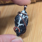 Hand holding a black and white stone pendant with copper accents on a wooden surface.