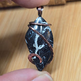 Hand holding a black and white marbled stone pendant with copper wire wrapping on a wooden surface.