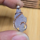 Close-up of a hand holding a purple stone pendant with a copper wire wrap design against a wooden background.