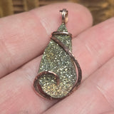 Teardrop-shaped pendant with glittery surface and copper wire wrapping held between fingers.