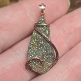 Teardrop-shaped pendant with glittery surface and copper wire wrapping held between fingers.