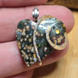 Heart-shaped stone pendant with silver and gold accents held between fingers.