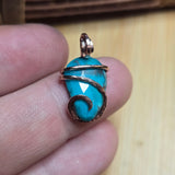 Turquoise stone pendant with rose gold filled wire wrapping held between fingers on a wooden surface