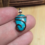 Turquoise stone pendant with rose gold filled wire wrapping held between fingers on a wooden surface