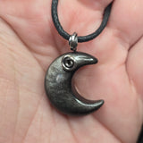 A close-up of a hand holding a dark gray crescent moon pendant necklace with a silver spiral accent and a black cord.