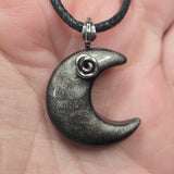 A close-up of a crescent moon pendant necklace resting on a person's hand. The pendant is dark gray with a metallic glitter finish and a small silver spiral detail near the top curve, hanging from a black cord.