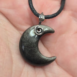 A close-up of a crescent moon pendant necklace resting on a person's palm. The pendant is dark gray with fine metallic flecks and a silver spiral accent near the top, hanging from a black cord.