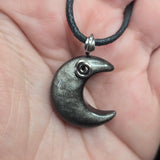 A close-up of a hand holding a crescent moon pendant necklace. The pendant is made of a dark, textured gray material with a small silver spiral detail near the top curve, suspended from a black cord.