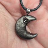 A close-up of a hand holding a crescent moon pendant necklace. The pendant is dark gray with a textured, metallic finish and a small silver spiral accent.