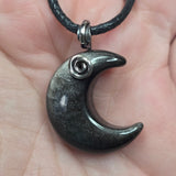 A close-up of a crescent moon pendant necklace held in a hand. The pendant is dark gray with fine metallic sparkle and a small silver spiral accent near the top, hanging from a black cord.