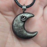 A close-up of a crescent moon pendant necklace held in a hand. The pendant is dark gray with a metallic glitter finish and a small silver spiral accent near the top curve, hanging from a black cord.