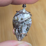 A close-up of a hand holding a leaf-shaped stone pendant wrapped with silver wire, set against a blurred wooden background.