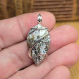 A raw mineral pendant wrapped in silver wire, held in a person's hand. The pendant features a natural, irregular shape with green, gray, and white crystalline textures, and a polished silver bail at the top.