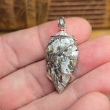 A teardrop-shaped polished stone pendant with a silver bail and silver wrap wire, held in a person's hand. The stone features a marbled pattern of white, gray, and brown.