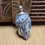 A leaf-shaped gemstone pendant with a silver wire-wrapped design, resting on a light wood surface in front of a brown leather strap.