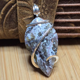 A leaf-shaped stone pendant with a silver bail, wrapped in silver wire, resting on a wooden surface with a brown leather strap in the background.