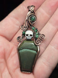 A gothic coffin-shaped pendant with a green stone body, a silver skull, and green gemstone accents, held in a person's hand.