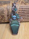A gothic-style coffin-shaped pendant featuring a dark green stone base, a silver skull detail, and a silver bail decorated with green gemstones and curled wire, displayed on a wooden surface with a leather book in the background.
