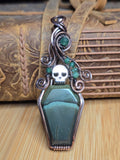 A gothic-style coffin-shaped pendant featuring a dark green stone body, a silver skull, and green gemstone accents, displayed against a rustic brown leather and wood background.