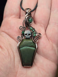 A gothic coffin-shaped pendant made of dark green stone and silver wire, held in a person's hand. The pendant features a small silver skull and green gemstone accents on a dark cord.