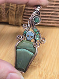 A gothic coffin-shaped pendant made of dark green stone and silver wire, held in a person's hand. The pendant features a small silver skull and green gemstone accents on a dark cord.