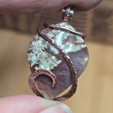 Close-up of a pendant with a natural stone and copper wire design held between fingers.