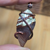 Hand holding a pendant with a stone and copper wire wrapping against a blurred background