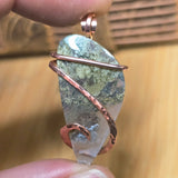 Pendant with a natural stone and copper wire wrapping held between fingers against a blurred wooden background