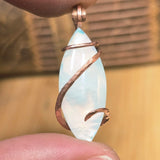 Marquis-shaped aurora opal pendant with copper wire wrapping held between fingers against a blurred background