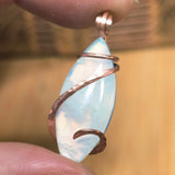 Marquis-shaped aurora opal pendant with copper wire wrapping held between fingers against a blurred background