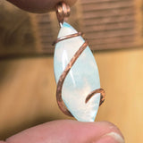 Pendant with a light blue stone and copper wire wrapping held between fingers against a blurred background