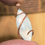 Pendant with a white stone and copper wire wrapping held between fingers against a blurred background