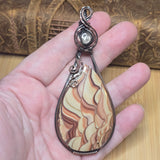 A hand holding a teardrop-shaped pendant featuring a marbled stone with cream, tan, and brown wavy patterns, wrapped in silver-toned metal with a small silver bead at the top.