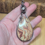 A teardrop-shaped pendant with a marbled agate-like pattern in cream, tan, and brown, wrapped in a silver-toned metal frame and held in a person's hand.