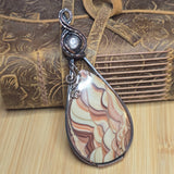 A teardrop-shaped pendant featuring a marbled agate or jasper stone in cream, tan, and brown tones, wrapped in dark silver-toned metal with decorative swirls, displayed in front of a brown leather journal on a bamboo surface.