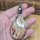 A hand holding a teardrop-shaped pendant made from a marbled agate or jasper stone in cream, tan, and brown tones, wrapped in silver-toned metal with a small dark bead at the top.