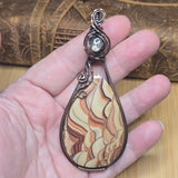 A hand holding a teardrop-shaped pendant made from a marbled beige, brown, and cream stone in a dark metal wire frame, with a silver-toned swirl top.