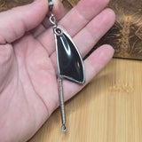 A hand holding a silver-toned pendant necklace featuring a large black  obsidian stone and a long coiled spring dangling from the bottom, with a small red gemstone accent at the top.
