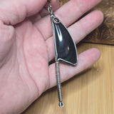 A hand holding a silver-toned pendant necklace featuring a polished black teardrop-shaped stone and a long coiled spring dangling from the bottom, set against a wooden surface.