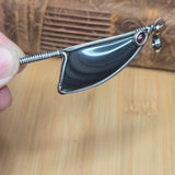A close-up of a handmade pendant featuring a polished black obsidian stone wrapped in silver wire, held by a hand over a light wood background.