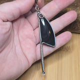 A hand holding a unique silver-toned pendant with a glossy black teardrop-shaped stone, a small red gemstone accent, and a coiled spring dangling from the bottom, set against a wooden background.