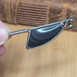 A handmade silver wire-wrapped pendant featuring a polished black sheen obsidian stone and a small red gemstone accent, held by a hand against a wooden background.