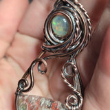 A close-up of a hand holding a handcrafted pendant featuring a round iridescent opal stone set in oxidized copper and silver wire, with a textured stone below.