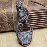 A handmade wire-wrapped pendant featuring a round iridescent opal and a larger shield-shaped stone with brown, white, and green crystalline patterns, set in dark silver-toned metal, displayed against a rustic brown leather and wood background.