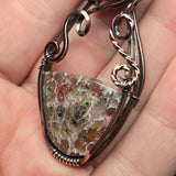 A close-up of a hand holding a handmade pendant made from a textured, mottled stone with a dark bronze wire wrap.