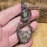 A hand holds a handmade wire-wrapped pendant featuring a round iridescent opal and a larger shield-shaped stone in a dark copper-toned metal setting, with a wooden box blurred in the background.