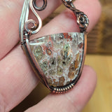 A close-up of a hand holding a handmade wire-wrapped pendant necklace. The pendant features a natural, irregularly shaped stone with a mottled pattern of brown, beige, and greenish tones, set in a copper-toned metal with a silver-toned chain.